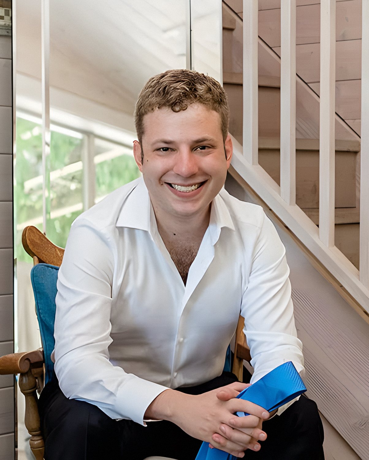 Mike Schneider in a crisp white shirt, sleeves rolled, grinning, hands clasped and leaning forward on a wooden chair.